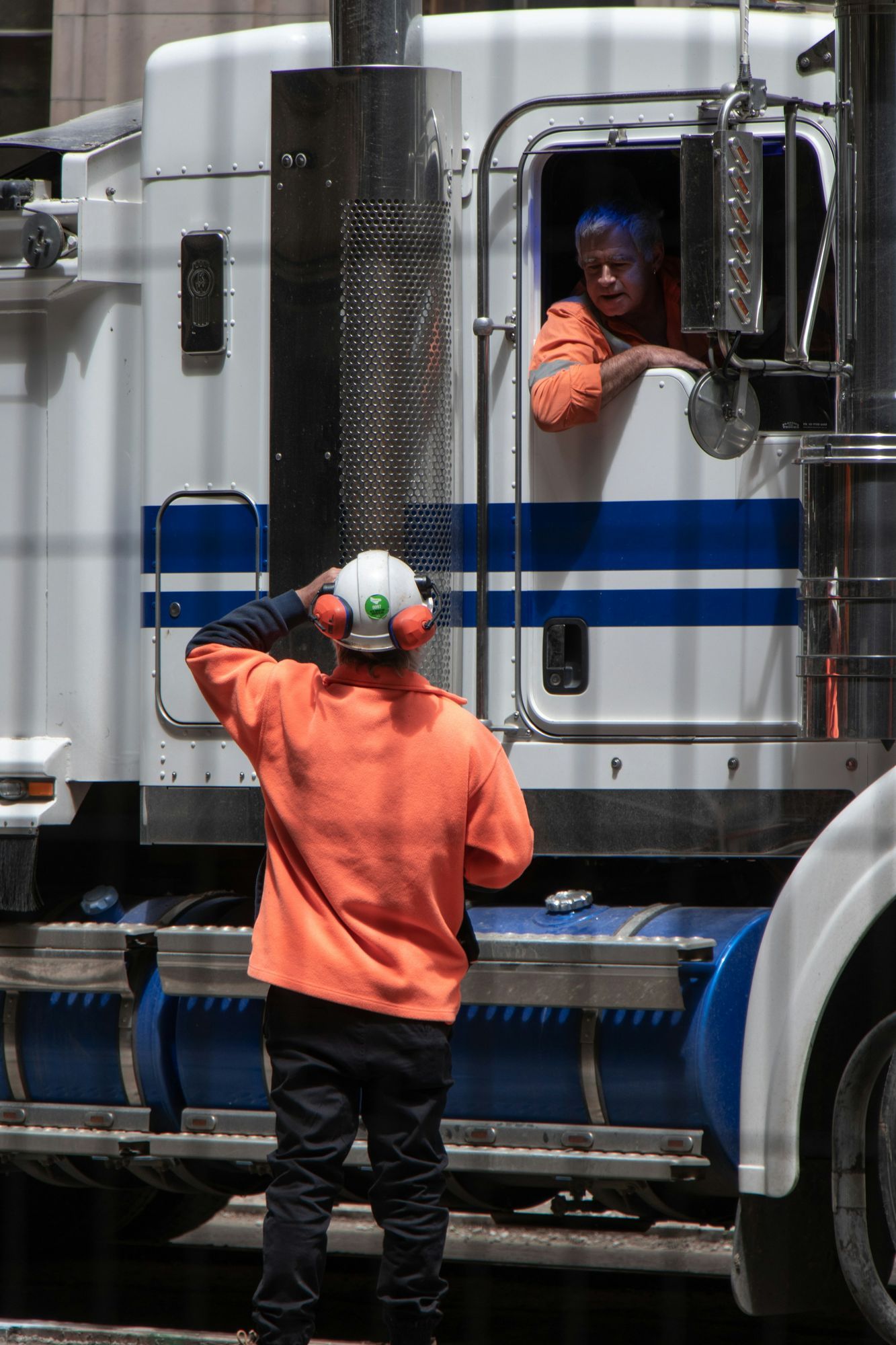 A worker in high-visibility clothing and a hard hat talking with a truck driver leaning out of the cab window.