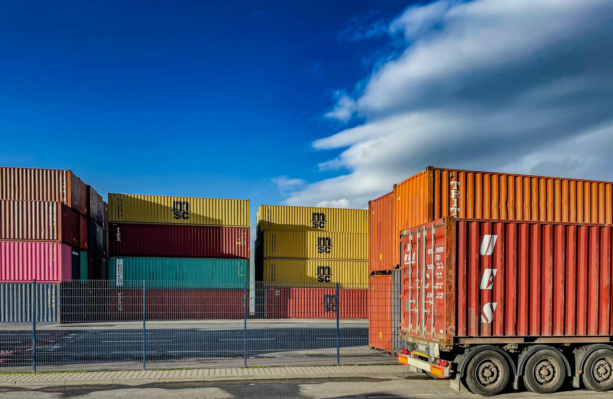 A loaded shipping container on a chassis beside stacked containers at a port terminal.