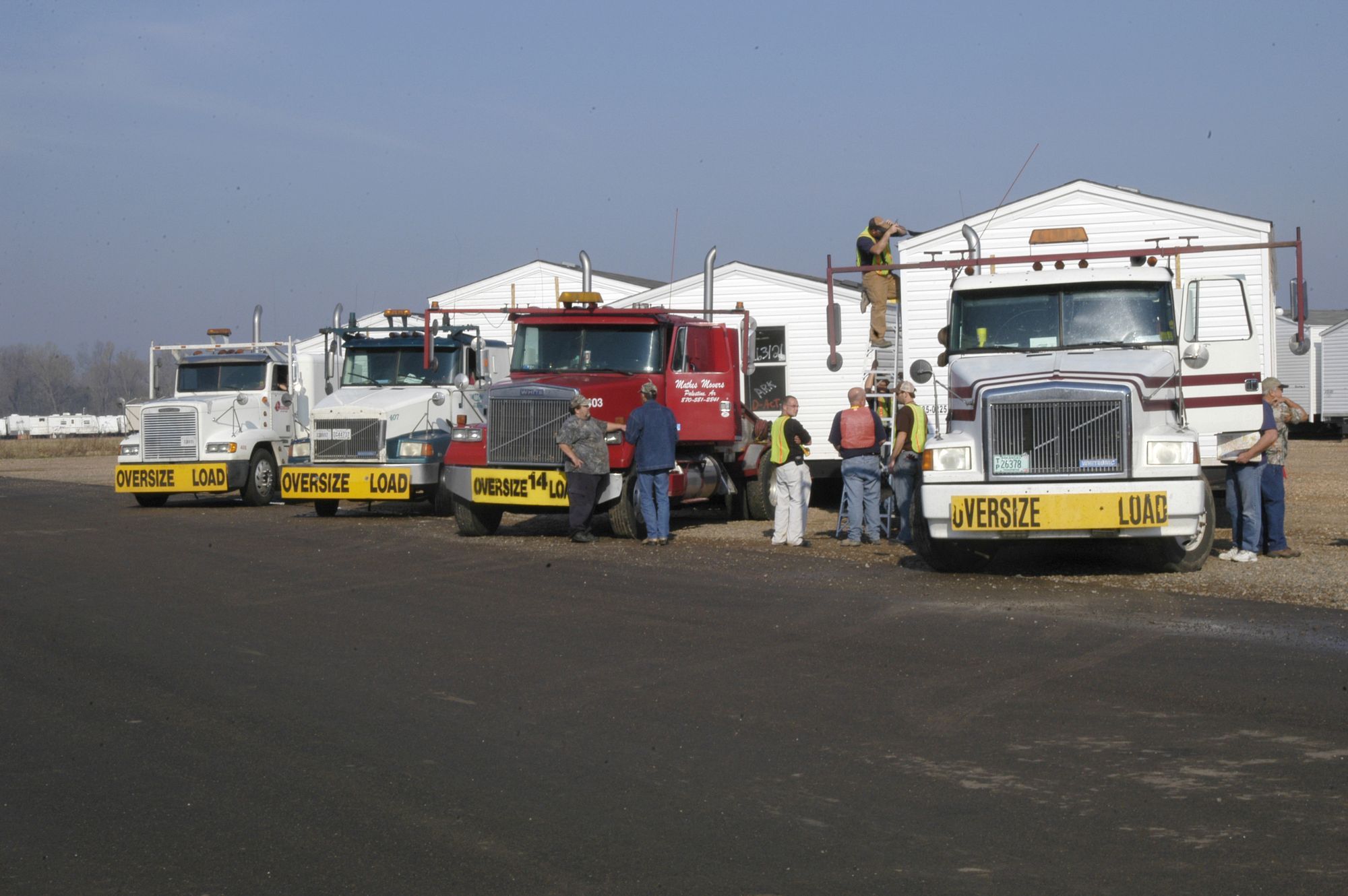 A line of toter trucks with OVERSIZE LOAD banners hitched to manufactured homes, crews preparing for departure.
