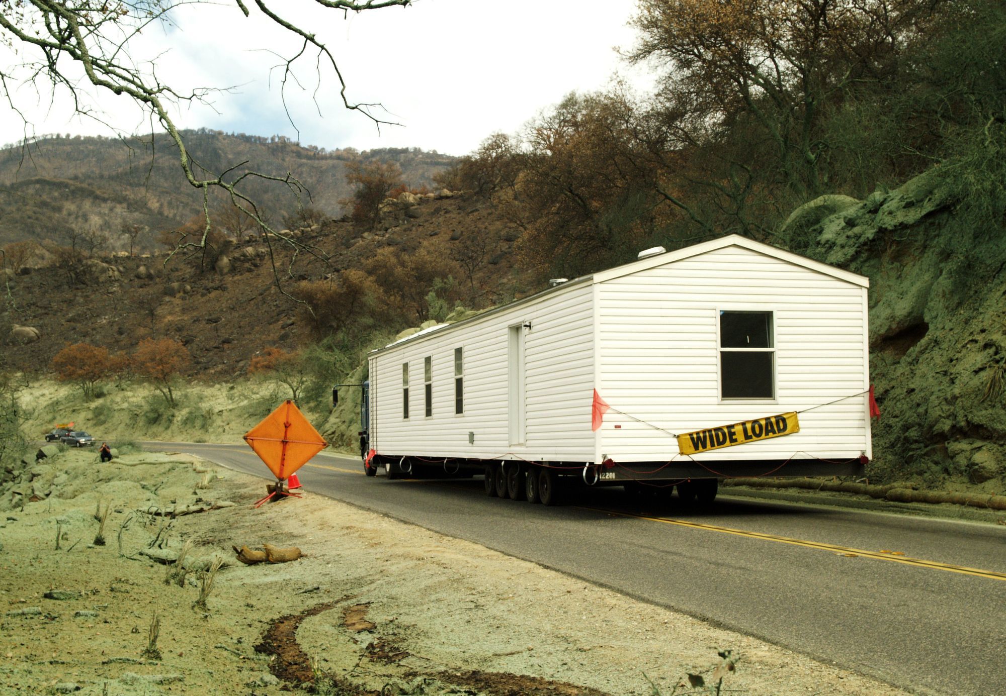 A single-section manufactured home with a WIDE LOAD banner being towed along a two-lane road.
