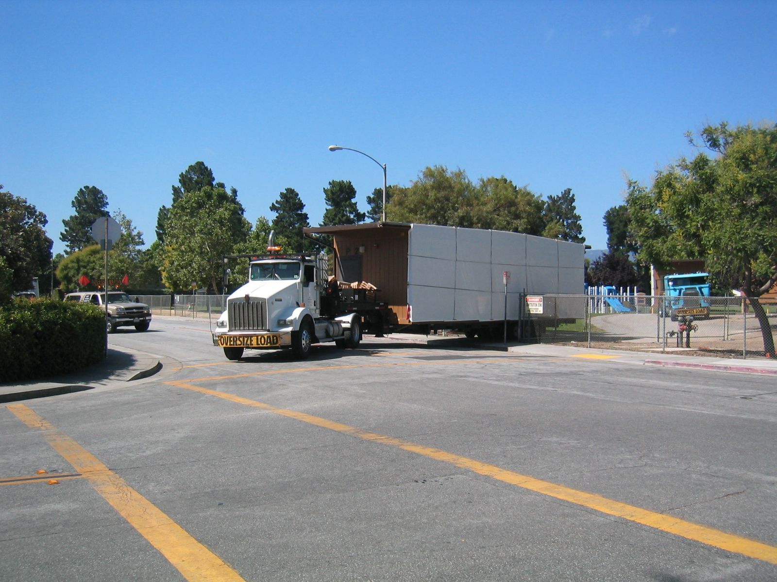 A white day-cab truck with an OVERSIZE LOAD banner moving a modular building section out of a site.
