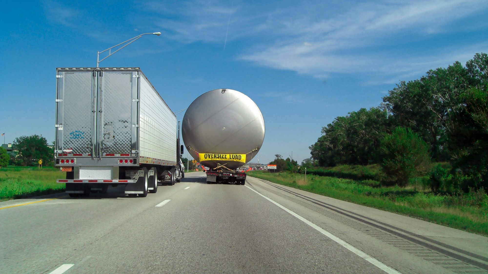 A large spherical tank carried on a trailer with an OVERSIZE LOAD banner, following a dry van down a highway.