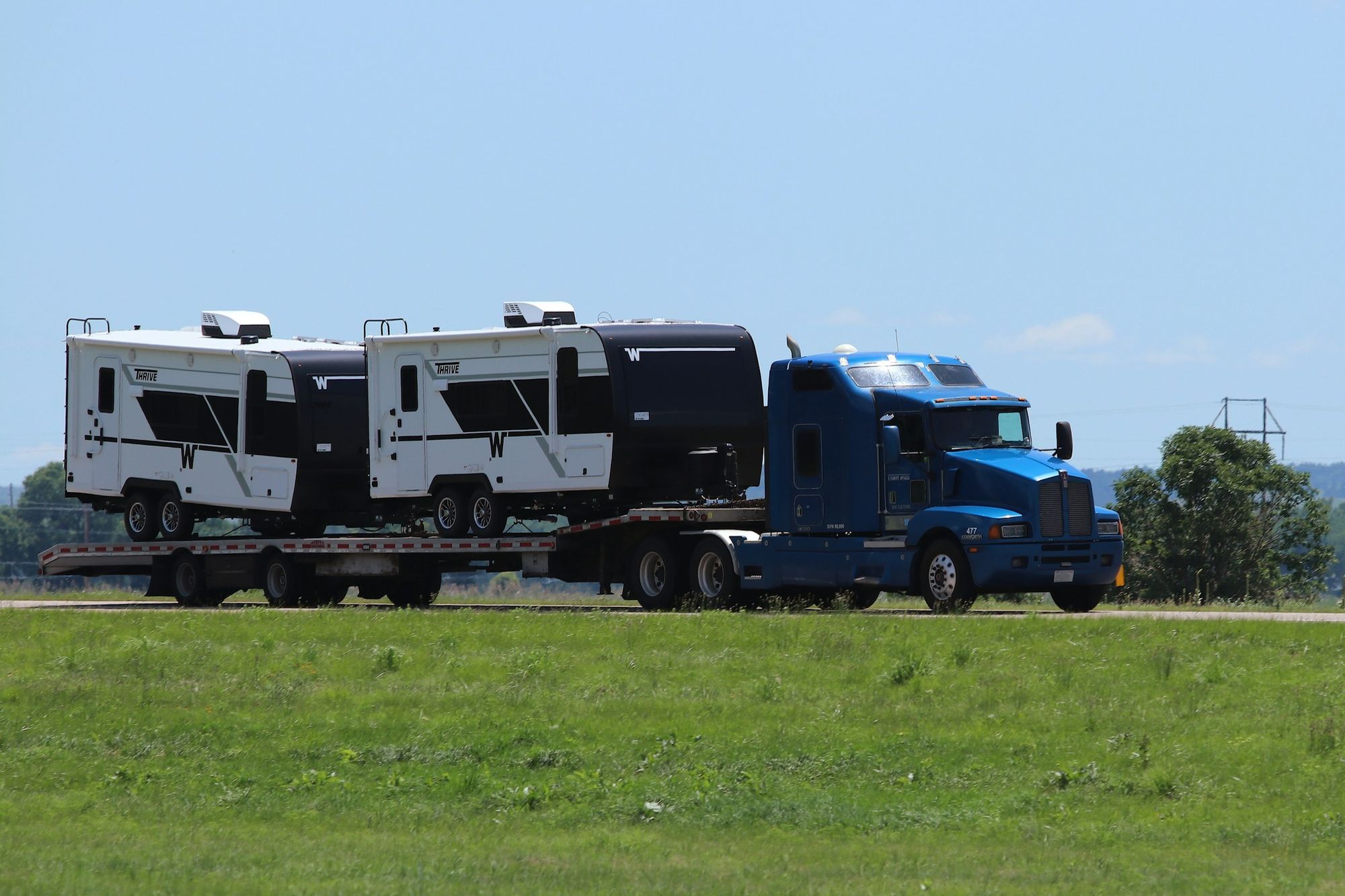 A blue semi-truck hauling two travel trailers on a flatbed trailer past a green field.